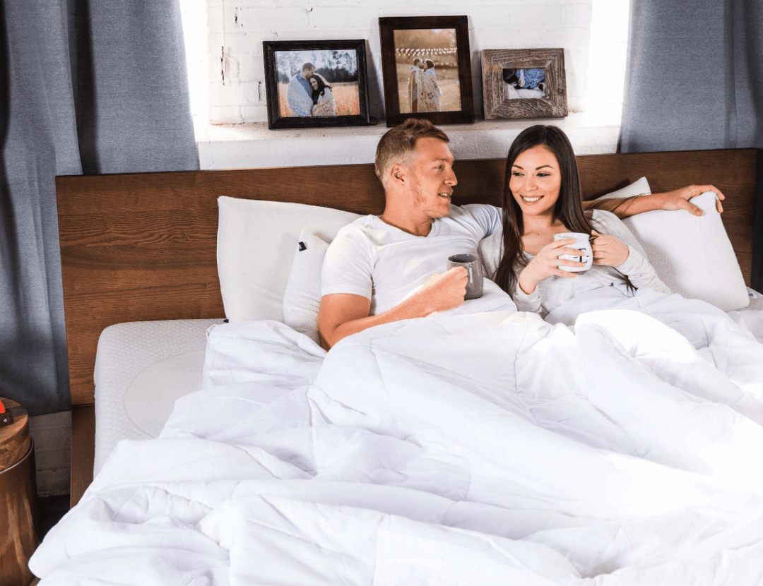 Couple sitting up in bed drinking coffee and smiling with white comforter, wooden headboard, and framed photos behind them
