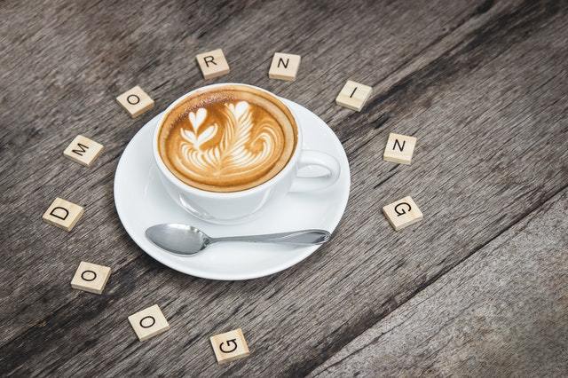 Cup of latte with heart-shaped latte art on a saucer with spoon, on a weathered wooden table surrounded by letter tiles spelling GOOD MORNING