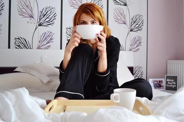 Woman in black pajamas sitting on a bed holding a large mug, breakfast tray on white bedding and floral wallpaper behind