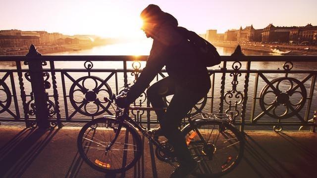 Cyclist riding a bicycle across a bridge at sunrise with a sunlit river and city skyline in the background