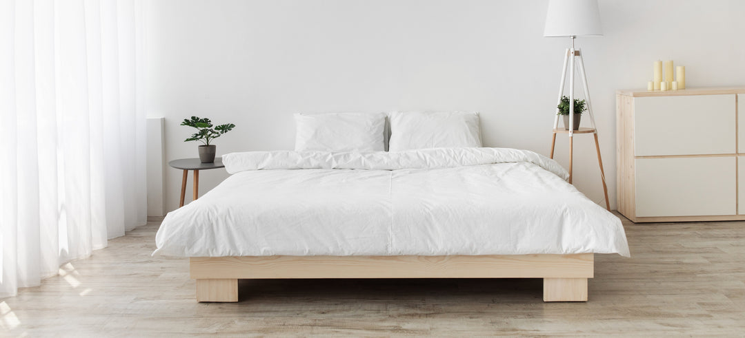 Bed centered in a bright minimalist bedroom with white linens on a low wooden platform, sheer curtains, a round nightstand with plant, floor lamp, and dresser
