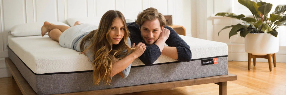 Smiling couple lying on their stomachs on a mattress on a wooden platform bed in a bright, plant-filled bedroom