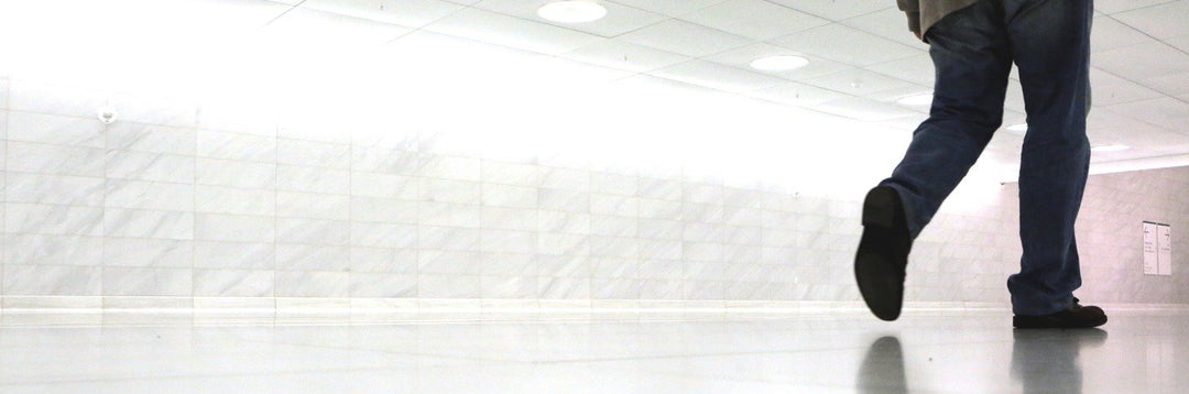 Person walking away in an empty, brightly lit tiled corridor showing lower legs and shoes reflected on the polished floor