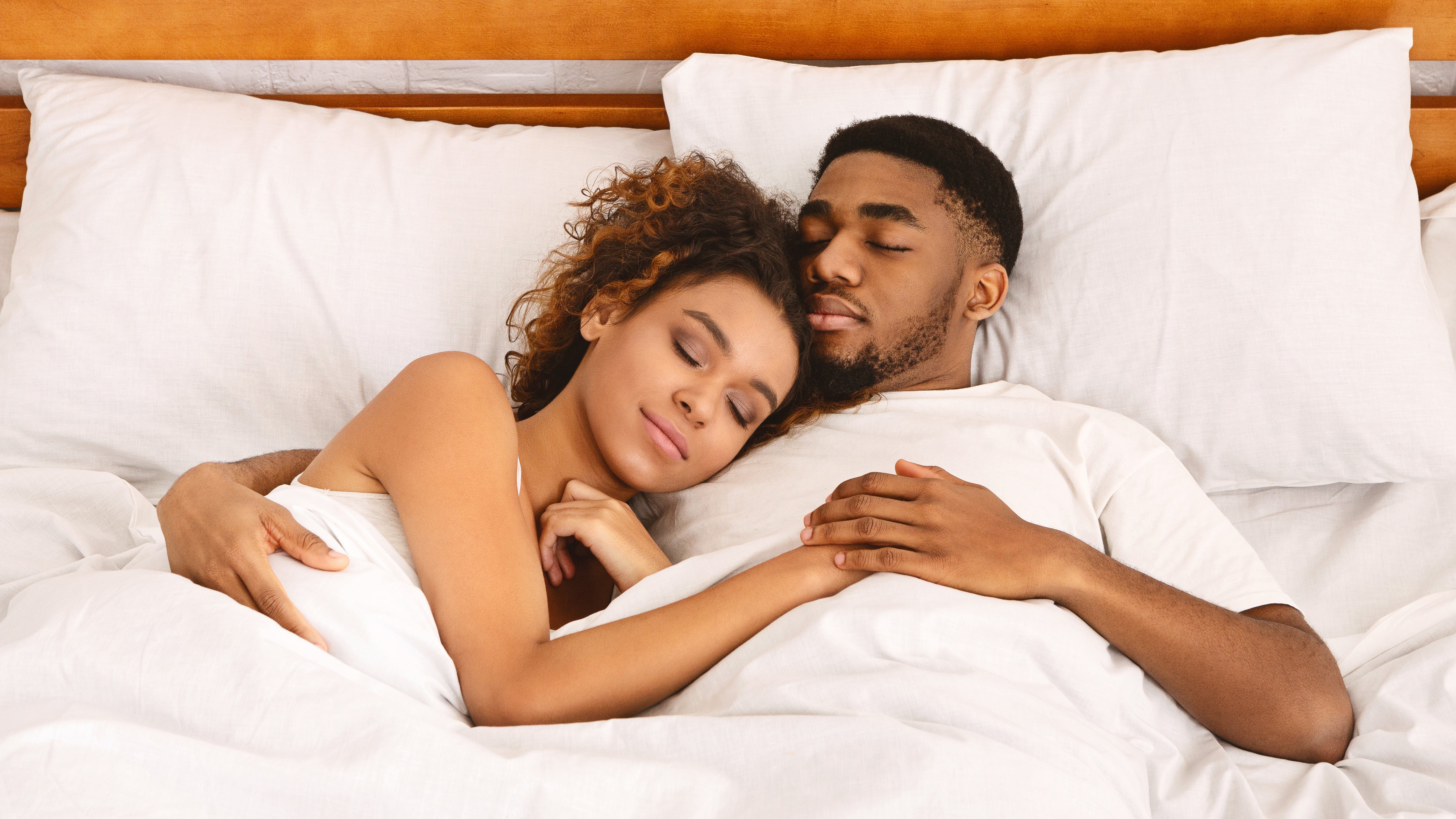 Couple cuddling and sleeping on a bed with white sheets and a wooden headboard