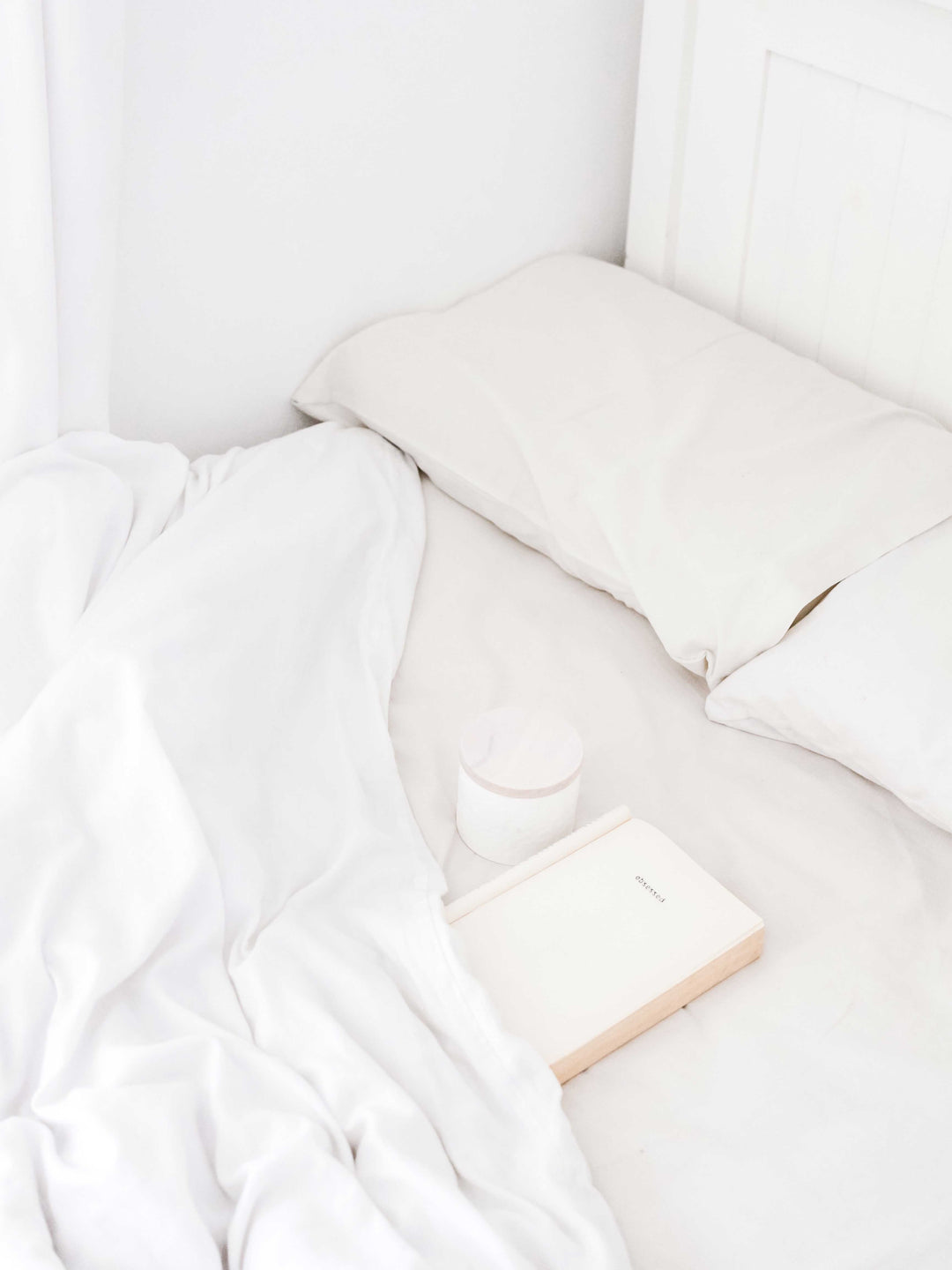 White full bed corner with pillow, rumpled sheets, and a closed journal on the mattress