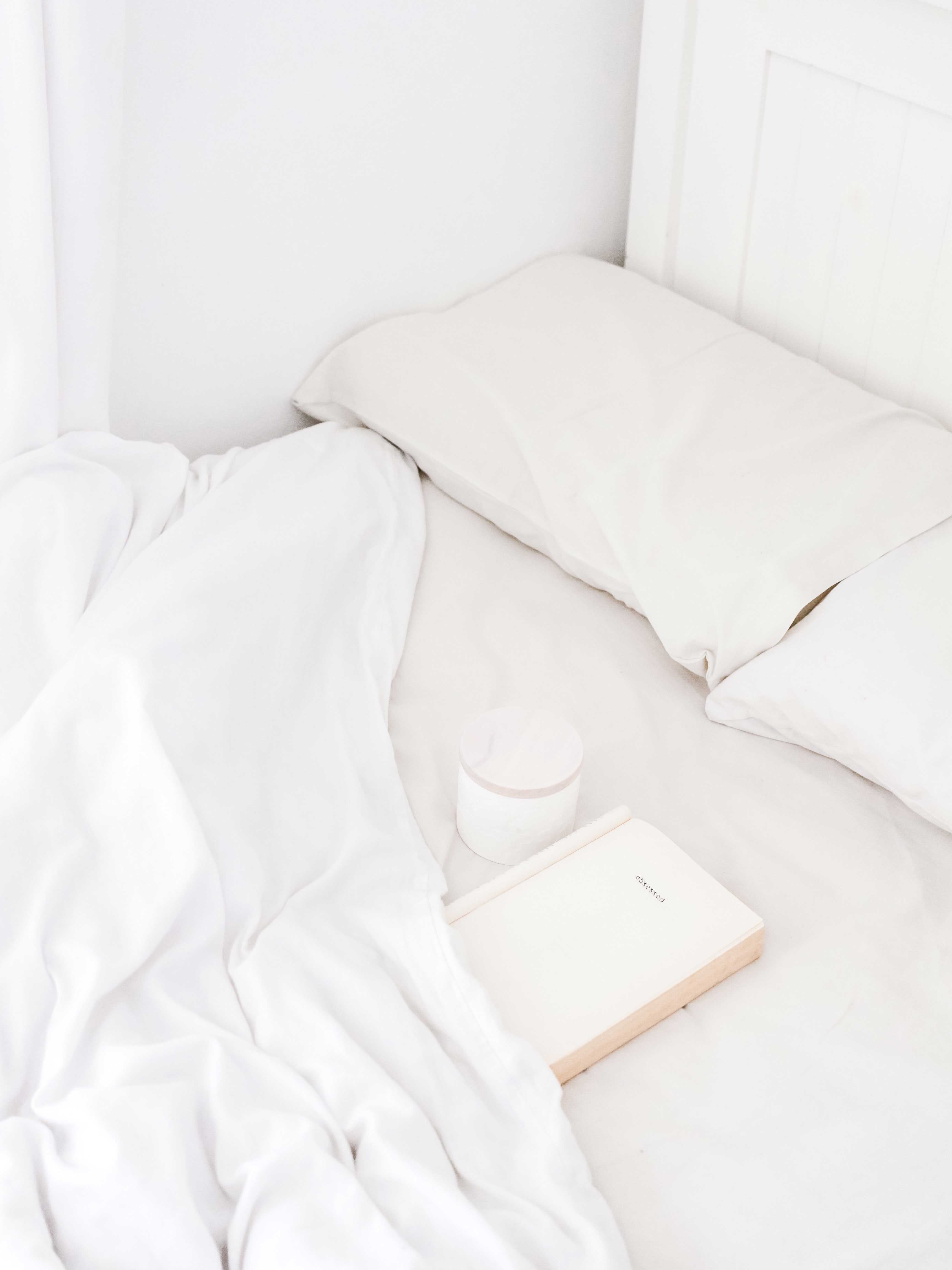 White full bed corner with pillow, rumpled sheets, and a closed journal on the mattress
