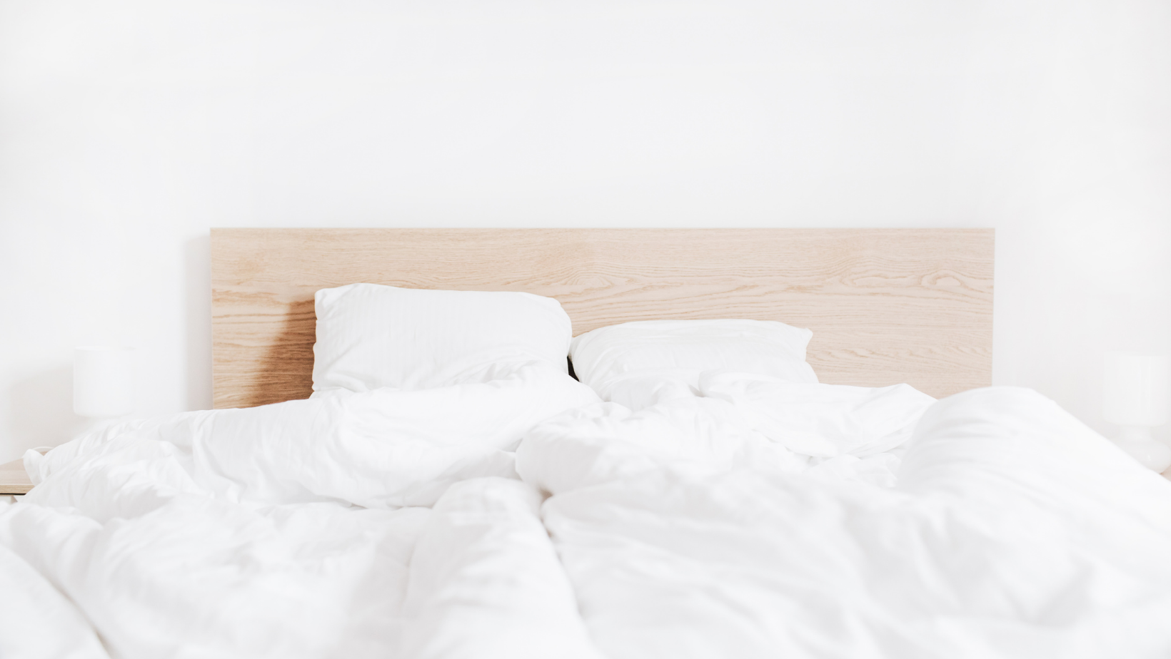 Unmade bed with white linens and pillows against a light wood headboard in a bright minimalist bedroom