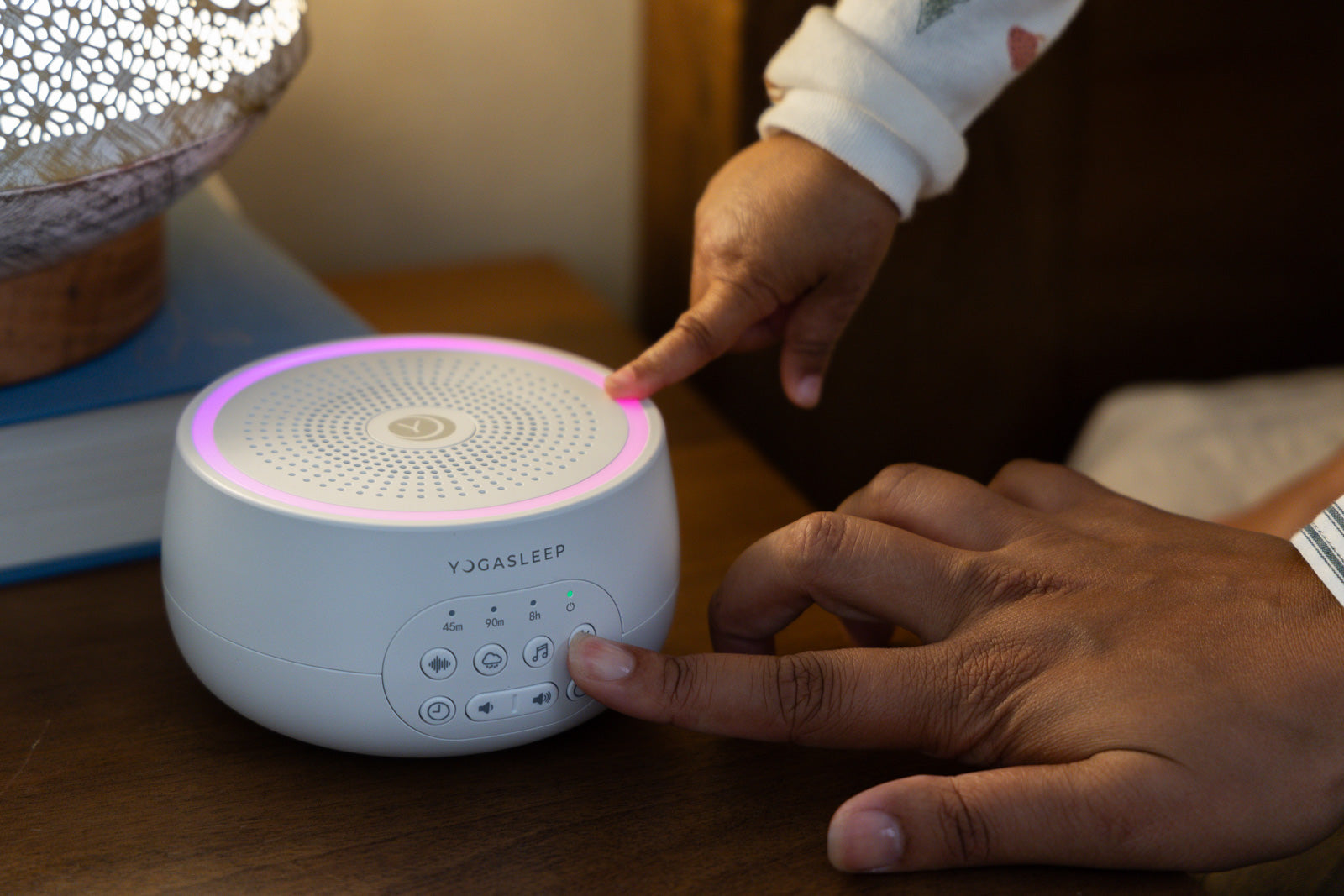 Top-front view of Yogasleep sound machine with pink LED ring and controls, on bedside table with adult and child hands