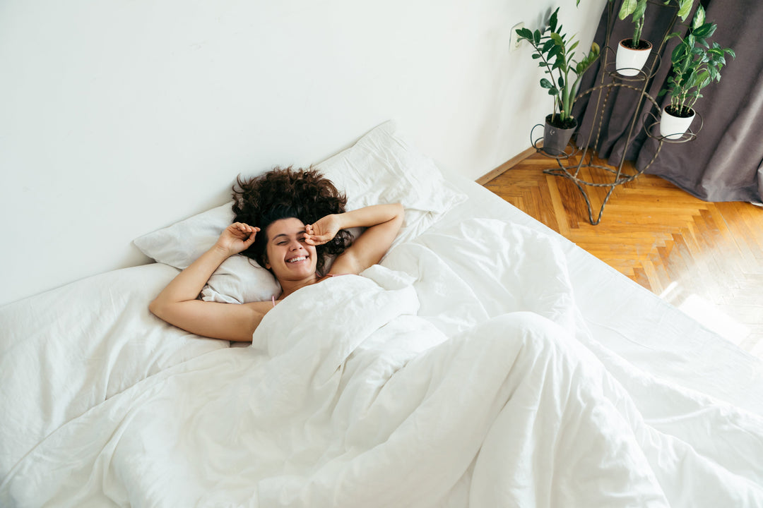 Smiling woman relaxing in a white bed in a bright bedroom with plants and hardwood floors