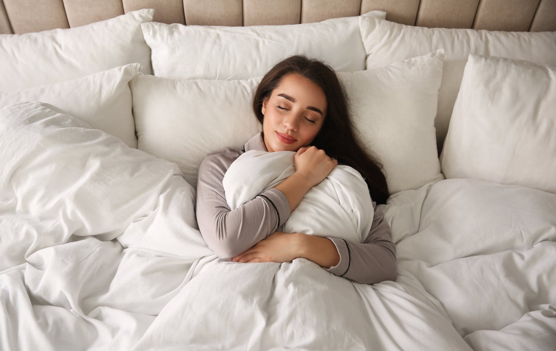 Woman hugging a pillow with eyes closed in bed surrounded by white pillows and bedding, looking peaceful