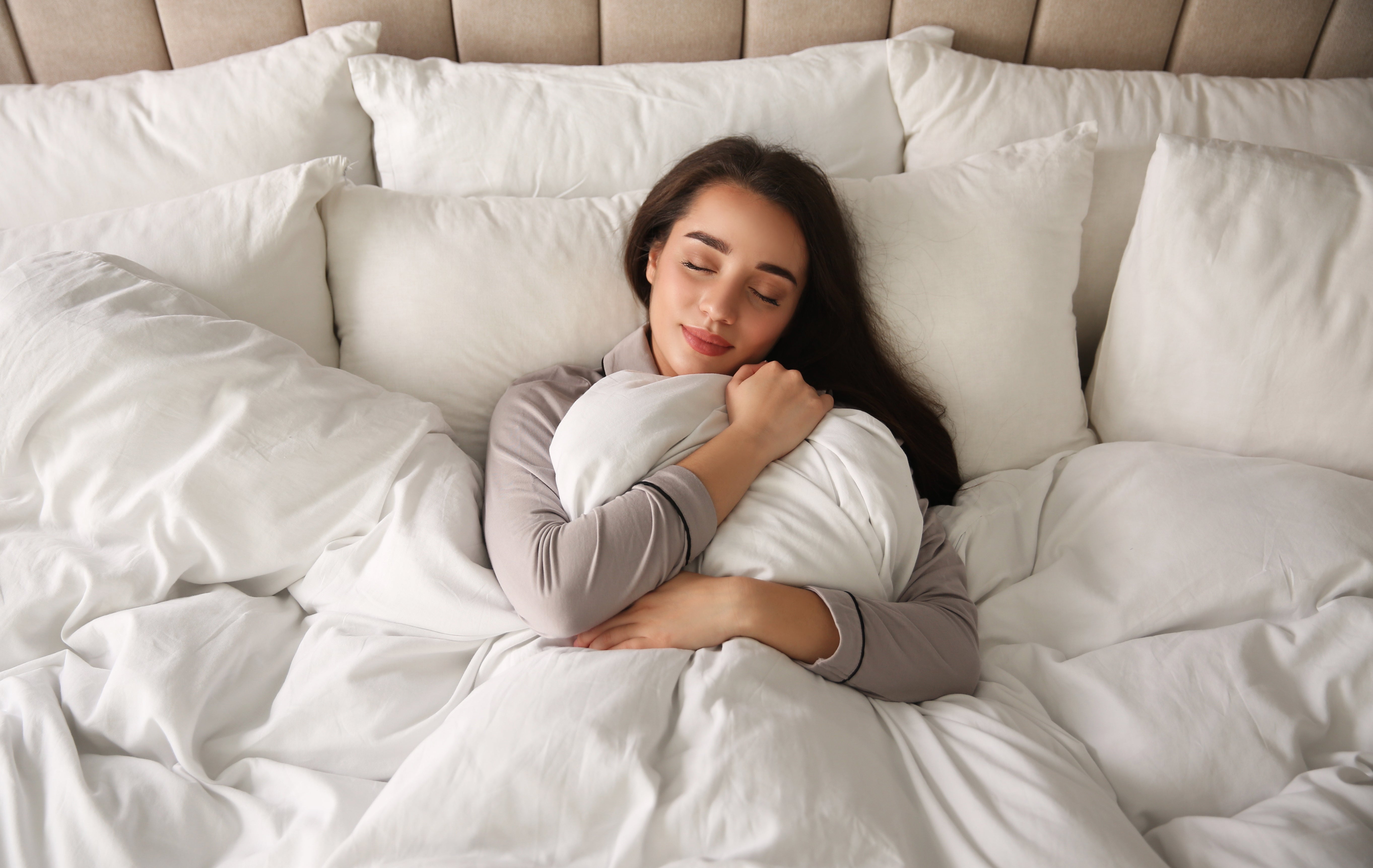 Woman hugging a pillow with eyes closed in bed surrounded by white pillows and bedding, looking peaceful