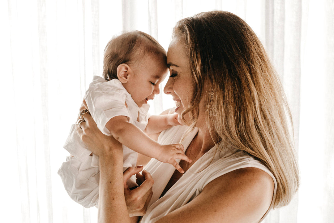 Mother holding and smiling at her baby, touching foreheads in a softly backlit room with sheer curtains