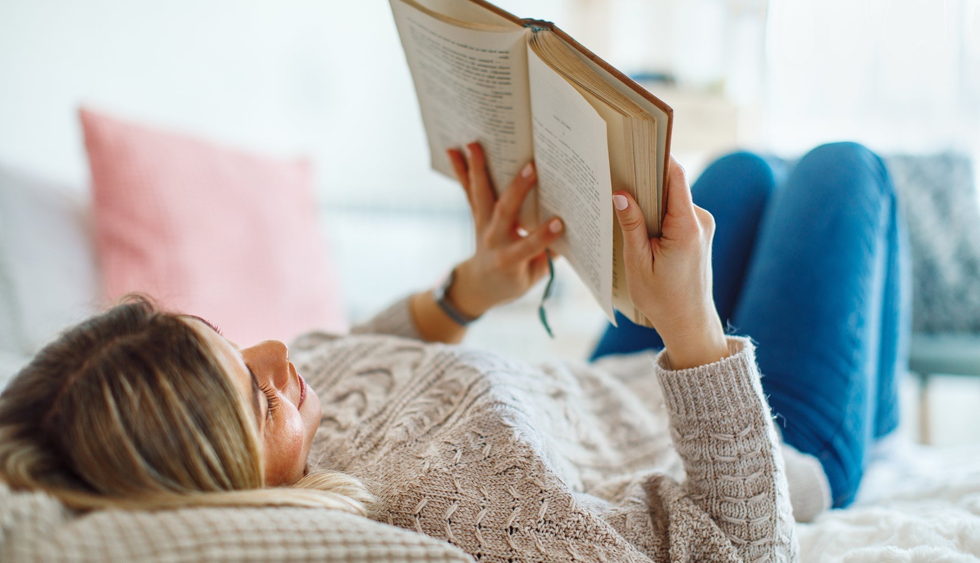 Woman lying on a bed reading a paperback book in a softly lit bedroom
