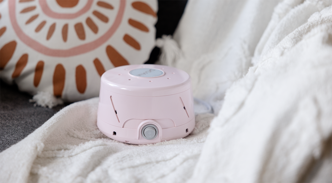 Angled front view of a pink white noise machine showing the front power dial and vent holes on a white blanket with a decorative pillow