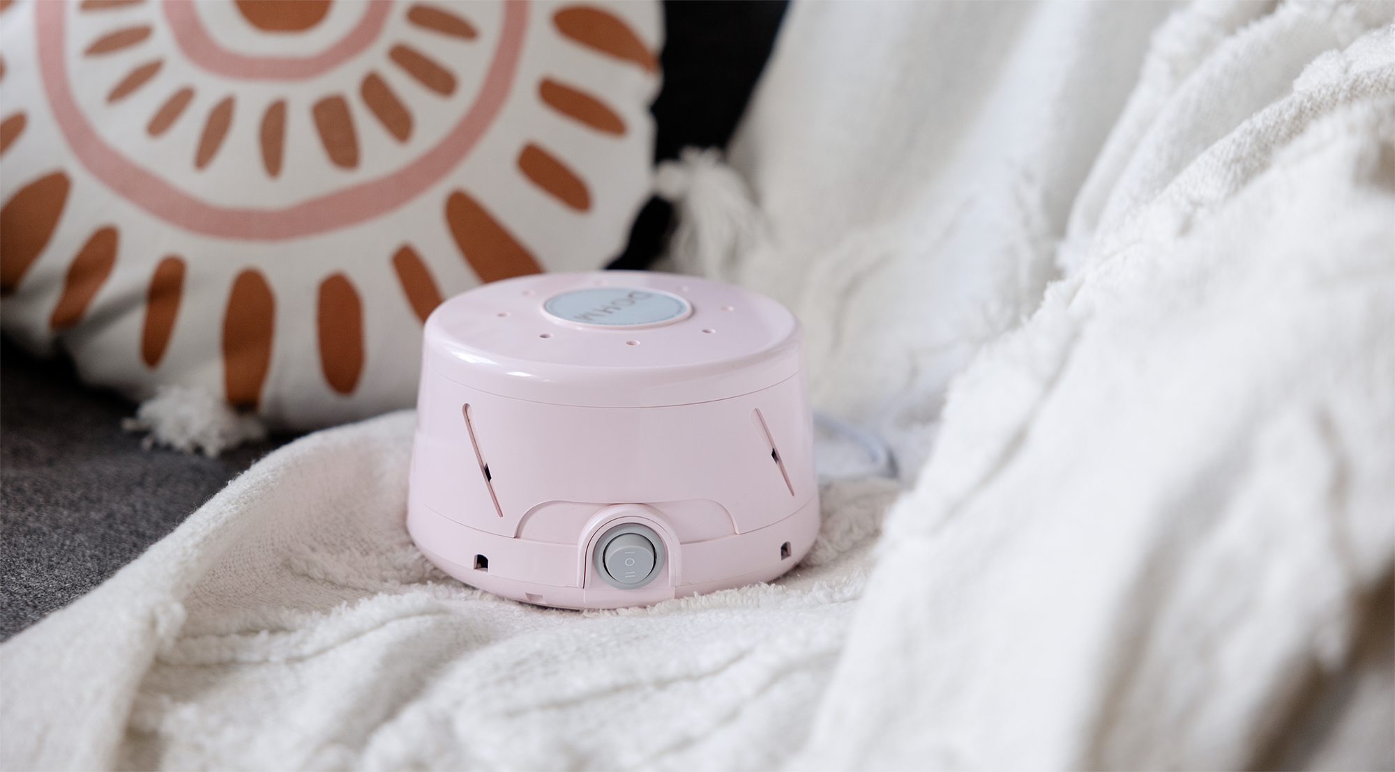 Angled front view of a pink white noise machine showing the front power dial and vent holes on a white blanket with a decorative pillow