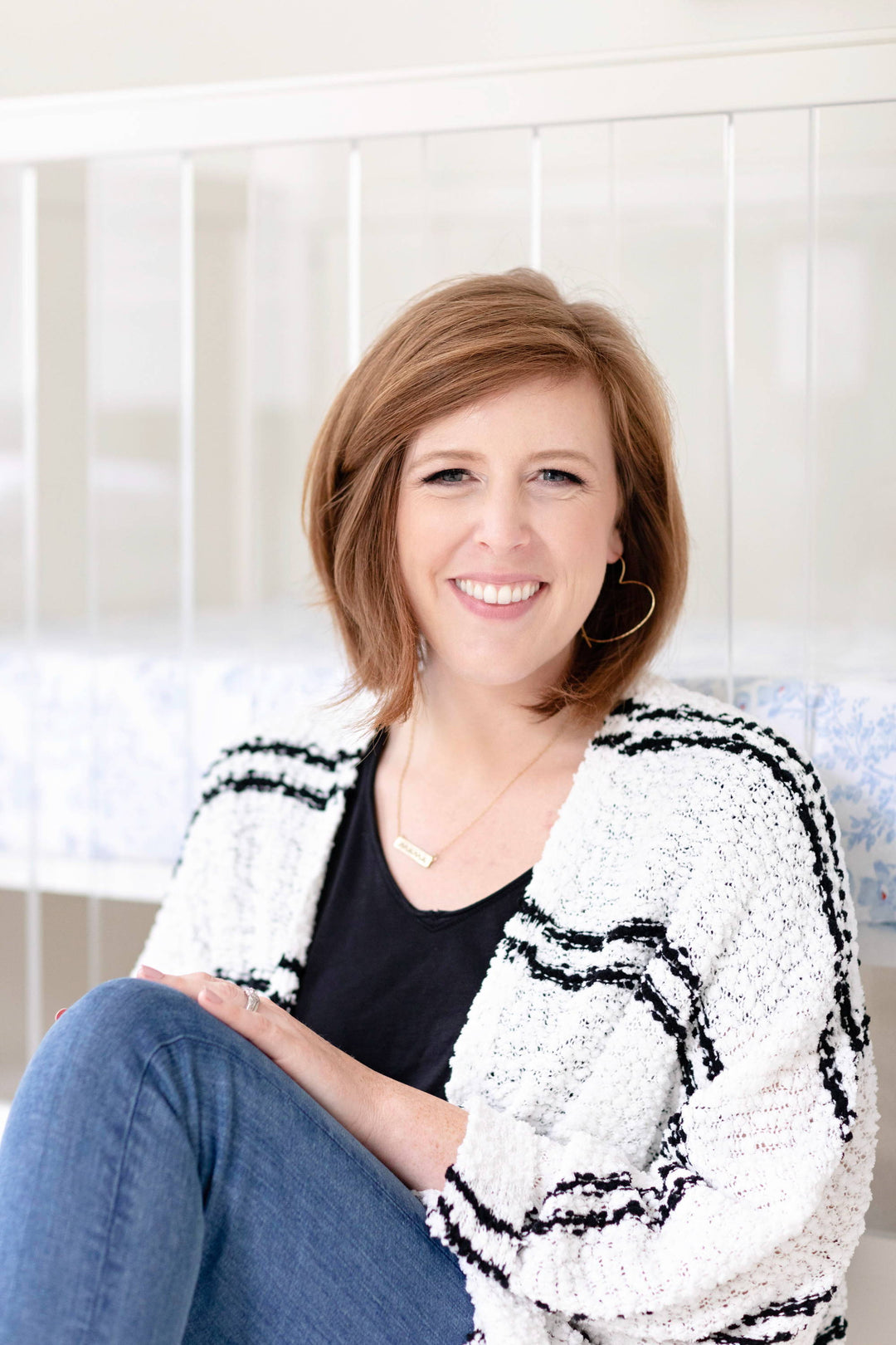 Smiling woman sitting in front of a white crib, wearing a black top and textured black-and-white cardigan