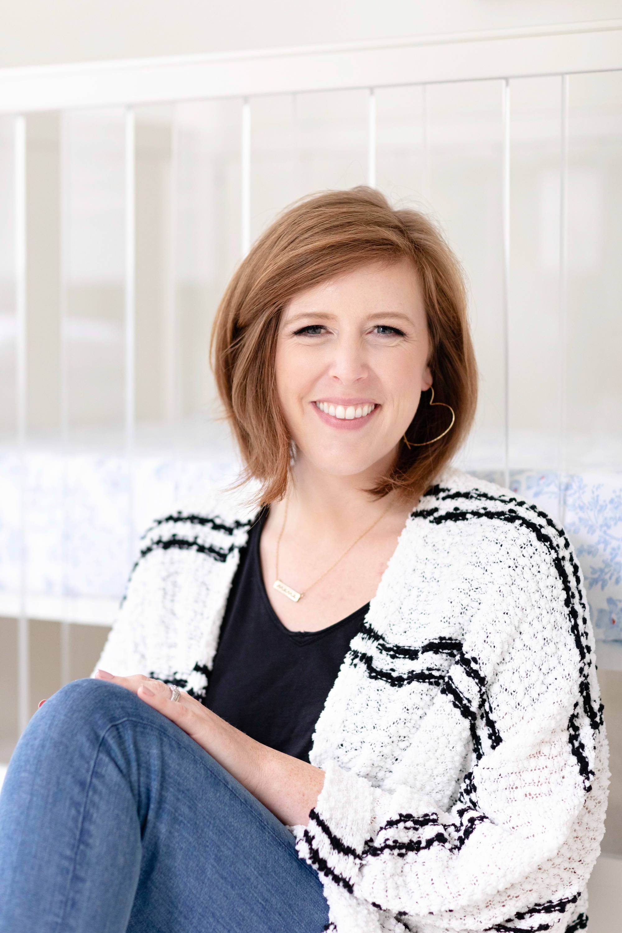 Smiling woman sitting in front of a white crib, wearing a black top and textured black-and-white cardigan