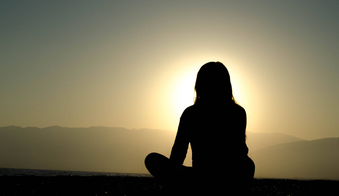 Silhouette of a person meditating cross‑legged on a beach at sunrise with distant mountains and calm sky