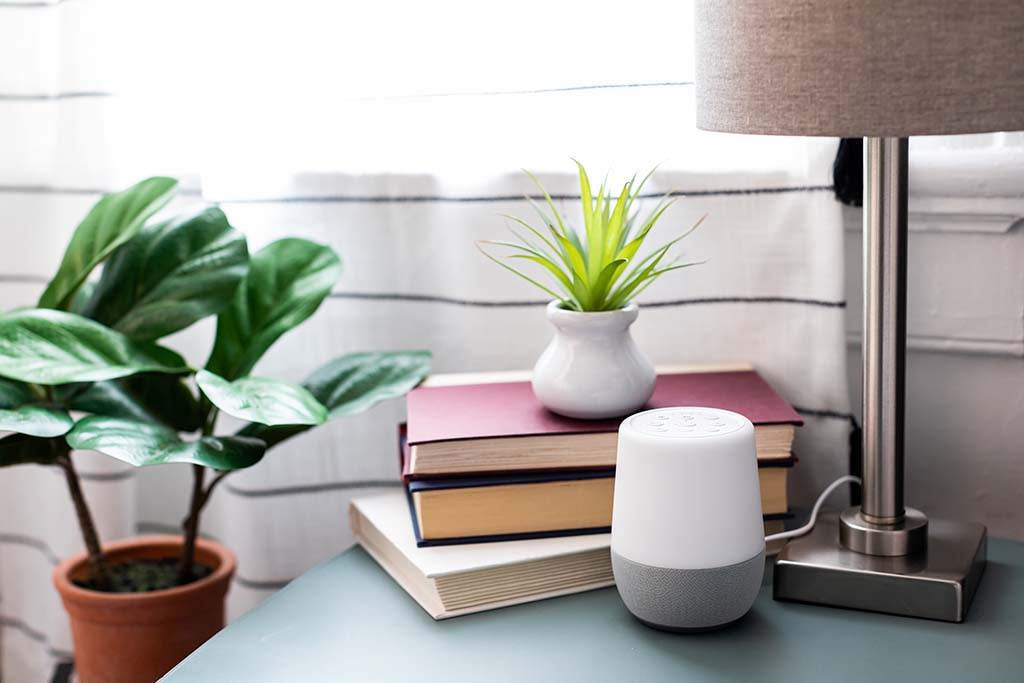 Front view of Yogasleep multi sound machine with white top and gray fabric base on a bedside table beside a lamp and potted plants