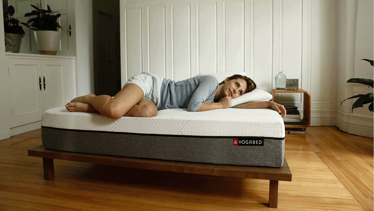 Side view of Yogabed mattress showing white textured top and gray sides on a wooden platform, woman lying on her side in a bright bedroom