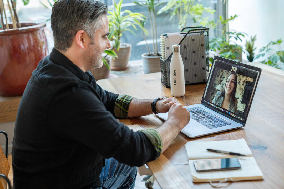Man on a video call at a wooden table, Postmates water bottle and notebook beside laptop in a plant-filled home office