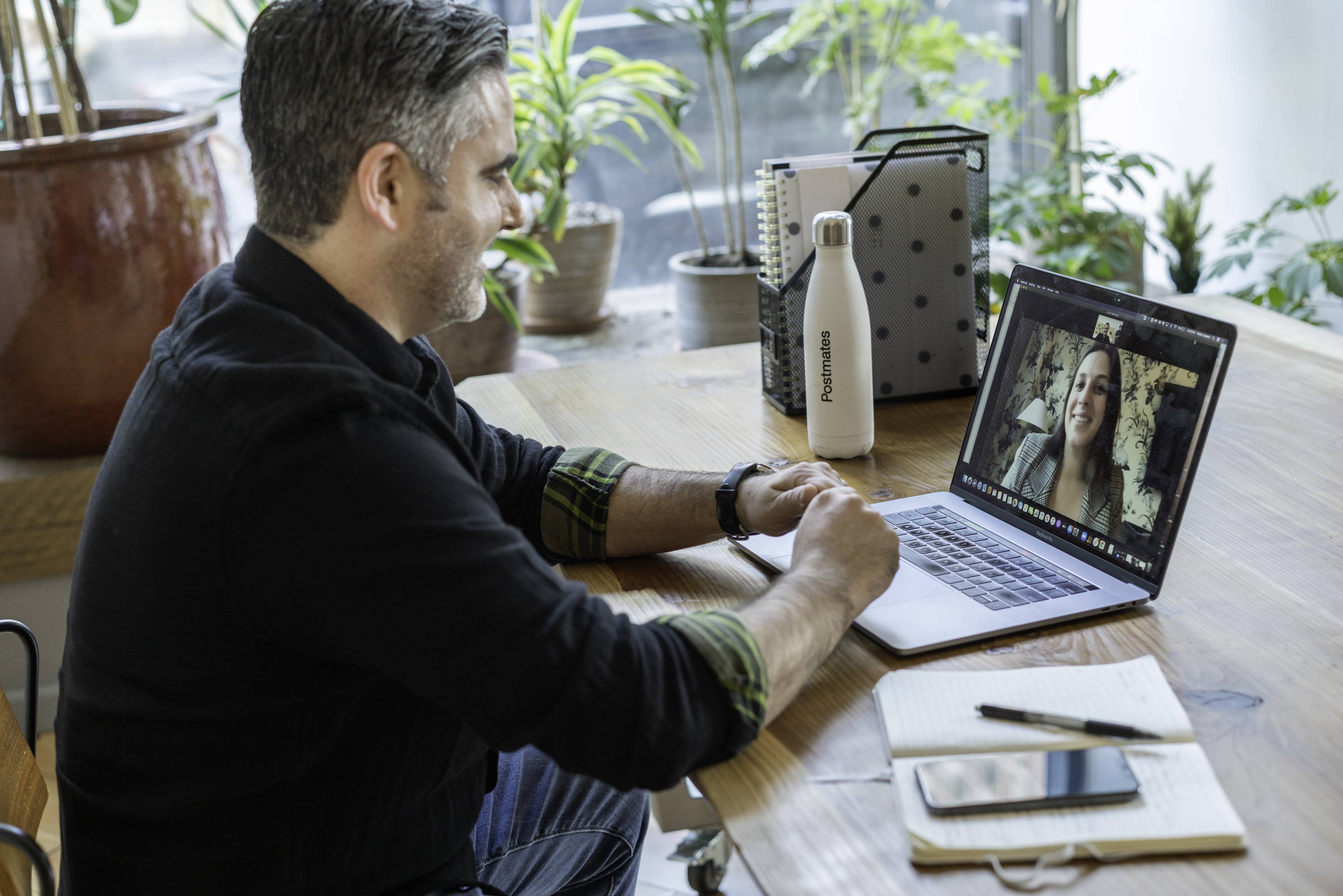 Man on a video call at a wooden table, Postmates water bottle and notebook beside laptop in a plant-filled home office