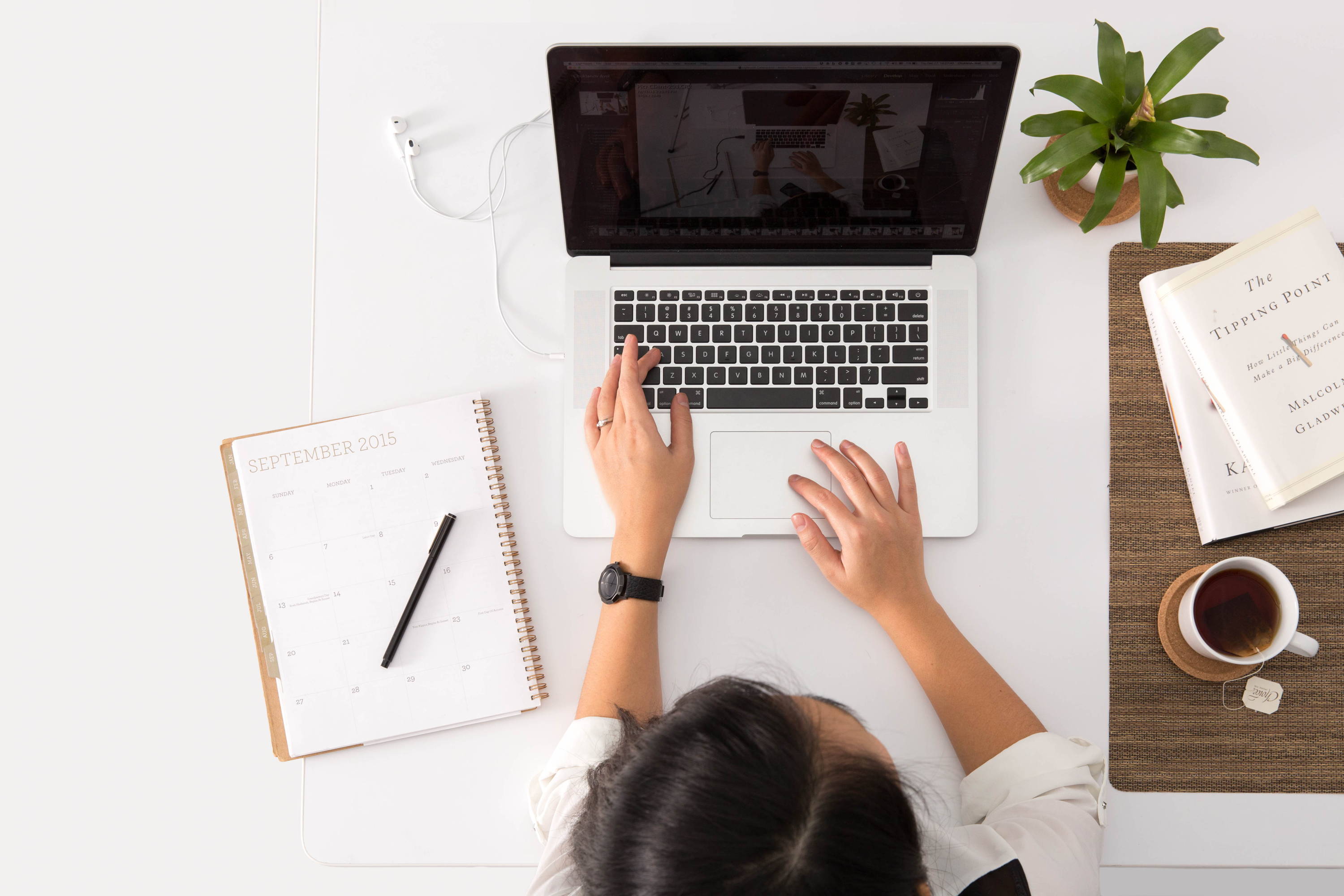 Overhead view of a person typing on a laptop at a tidy white desk with a planner, earbuds, potted plant, book, and cup of tea