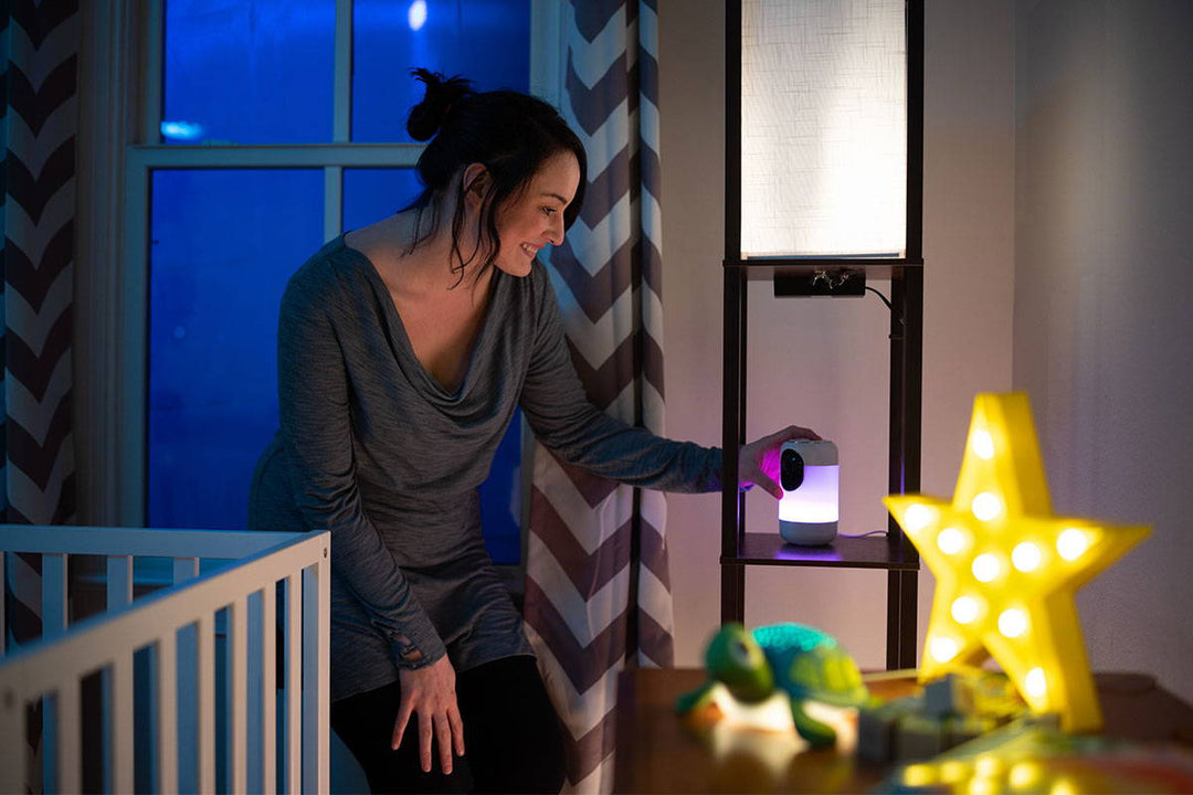 Mother adjusting a white noise machine on a nursery shelf beside a crib in a dim room with a star nightlight and turtle toy