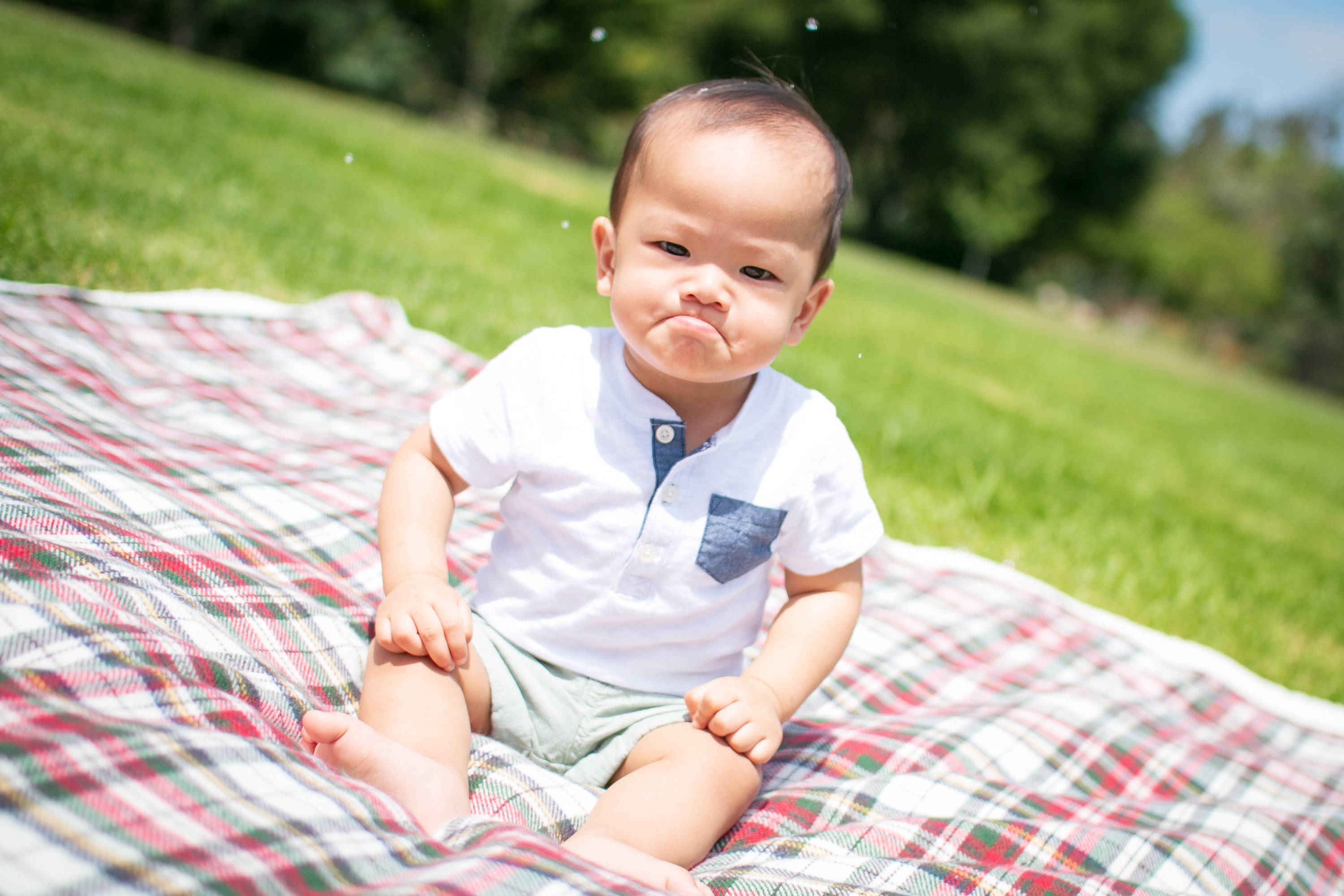 Baby sitting on a plaid picnic blanket in a sunny park making a pouty face