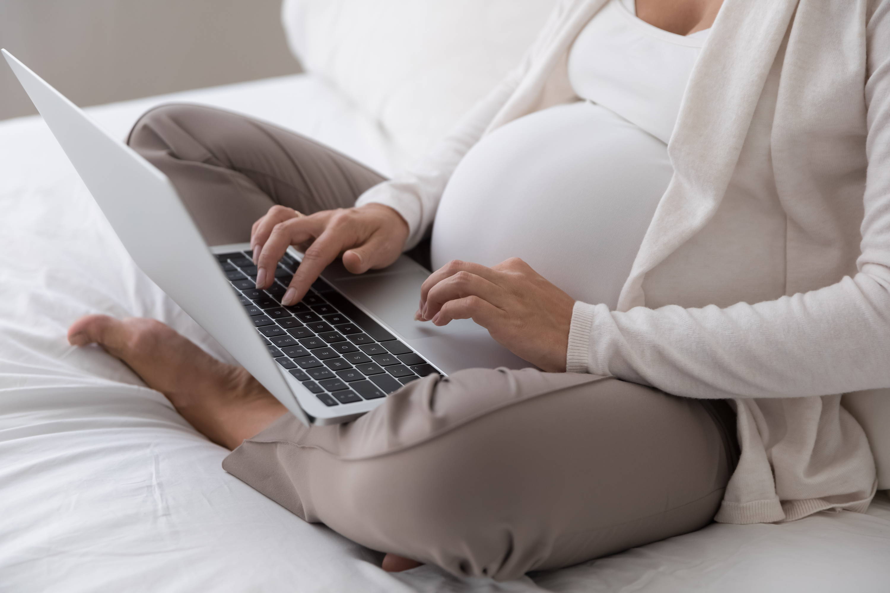 Pregnant person typing on a laptop while sitting cross-legged on a bed