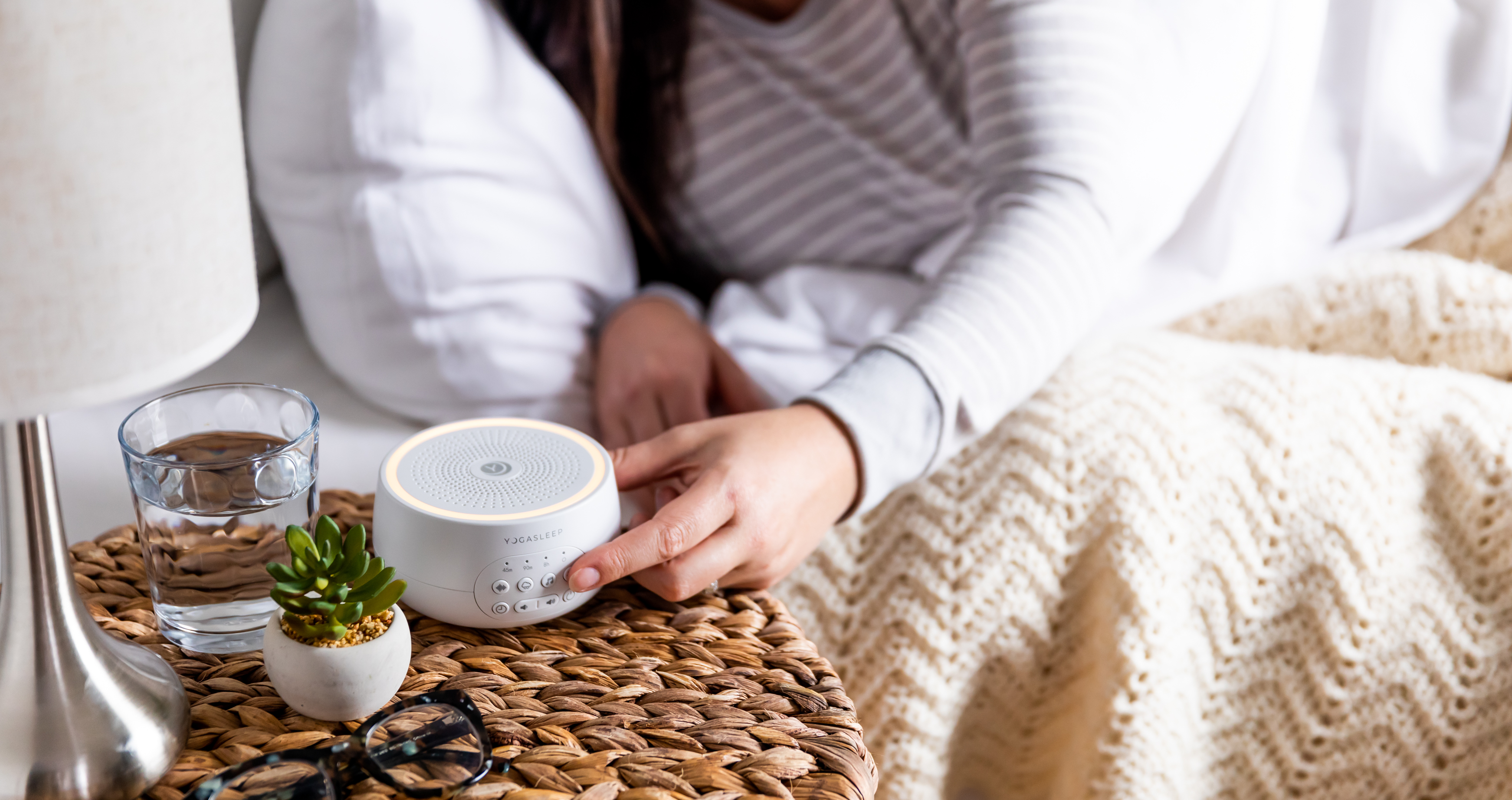 Person reaching to press a white Yogasleep sound machine on a bedside wicker table with a glass of water, lamp, and knitted blanket