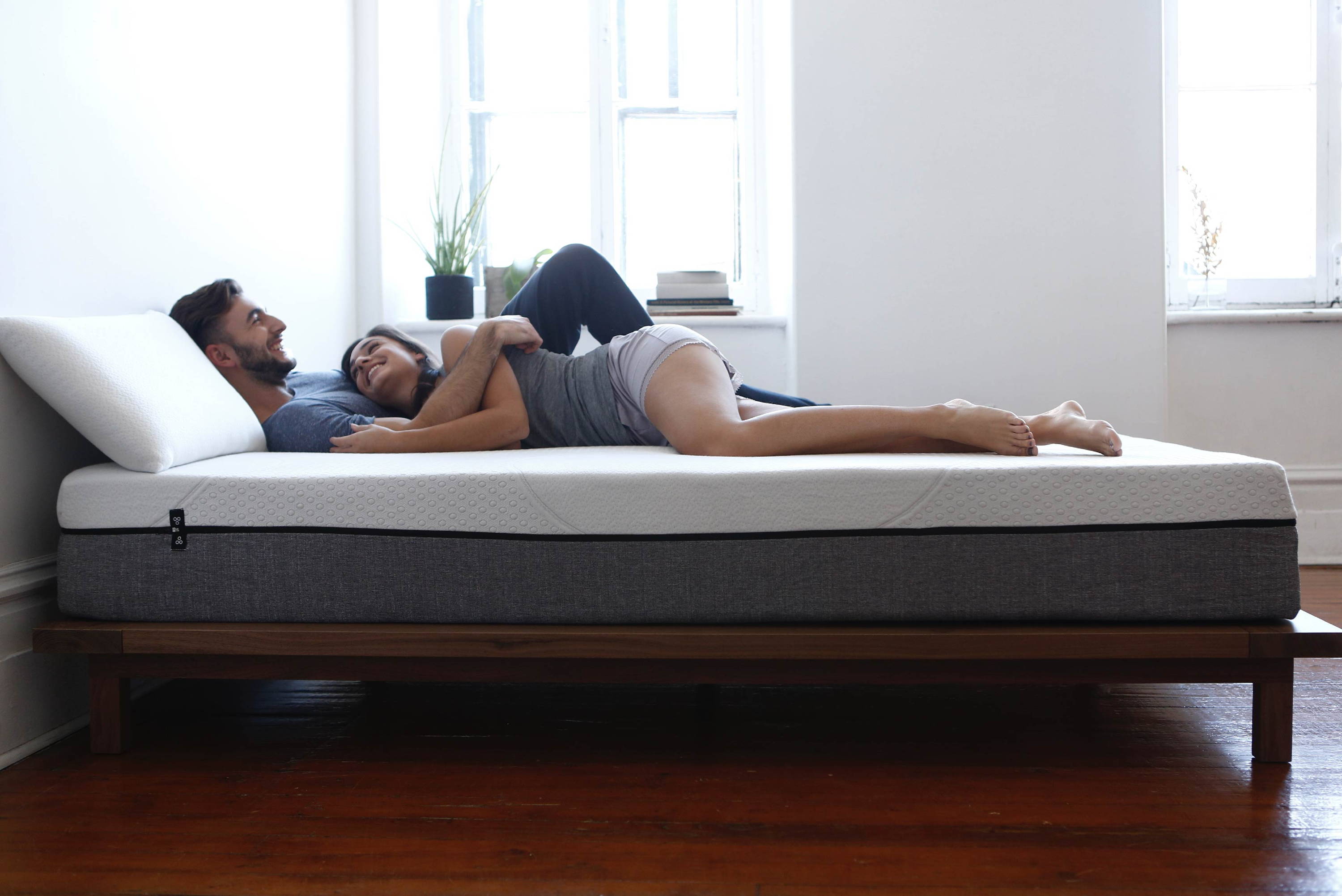 Couple lying and smiling on a white mattress on a low wooden platform bed in a bright bedroom with morning light