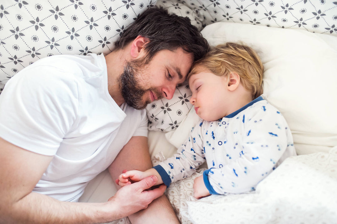 Father lying beside a sleeping toddler, holding the child's hand in bed