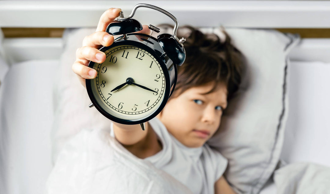 Child lying in bed holding a black twin-bell alarm clock up to the camera in a bedroom