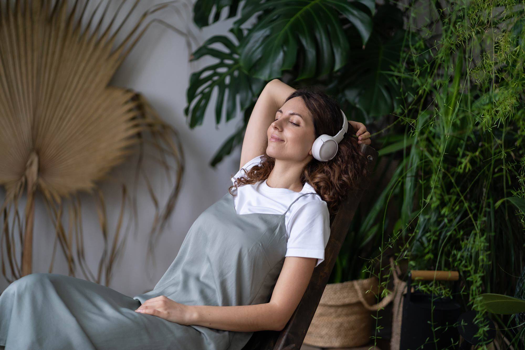 Woman wearing white headphones reclining with eyes closed among lush houseplants