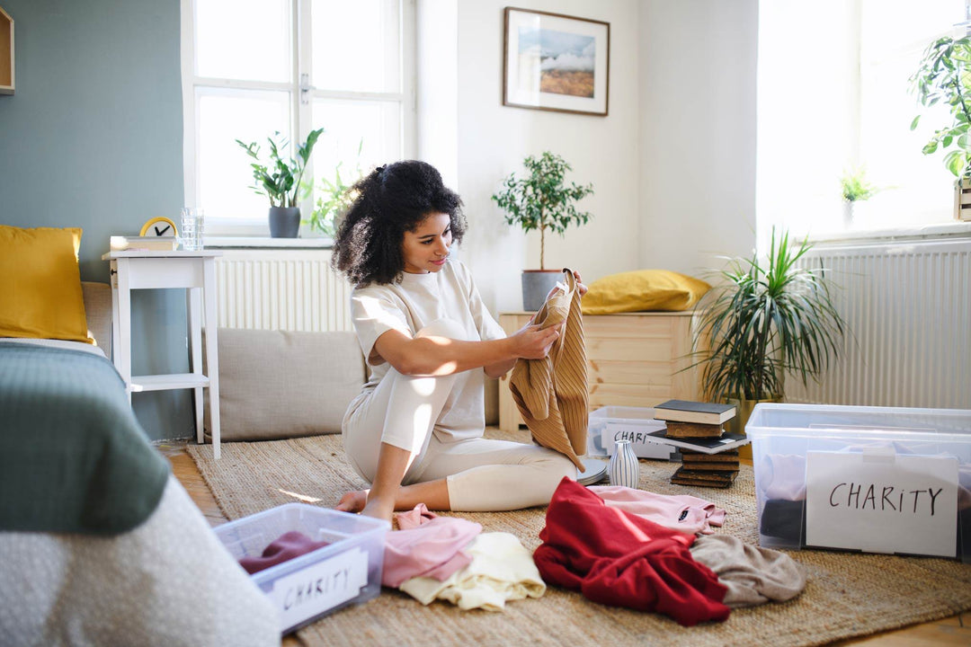 Woman sitting on a living room floor sorting clothes into plastic boxes labeled 'Charity' in a bright, plant-filled room