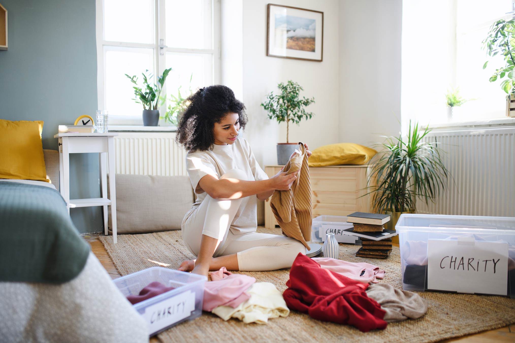 Woman sitting on a living room floor sorting clothes into plastic boxes labeled 'Charity' in a bright, plant-filled room
