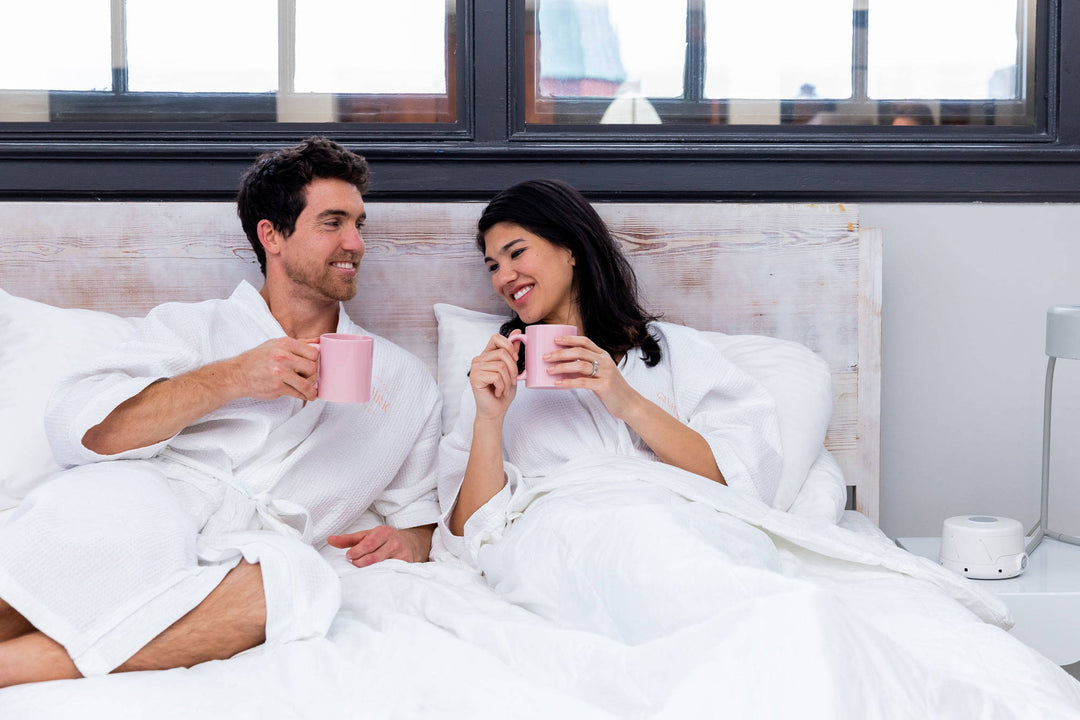 Man and woman in robes sharing coffee in bed with a white noise machine on the bedside table in a bright hotel-style room