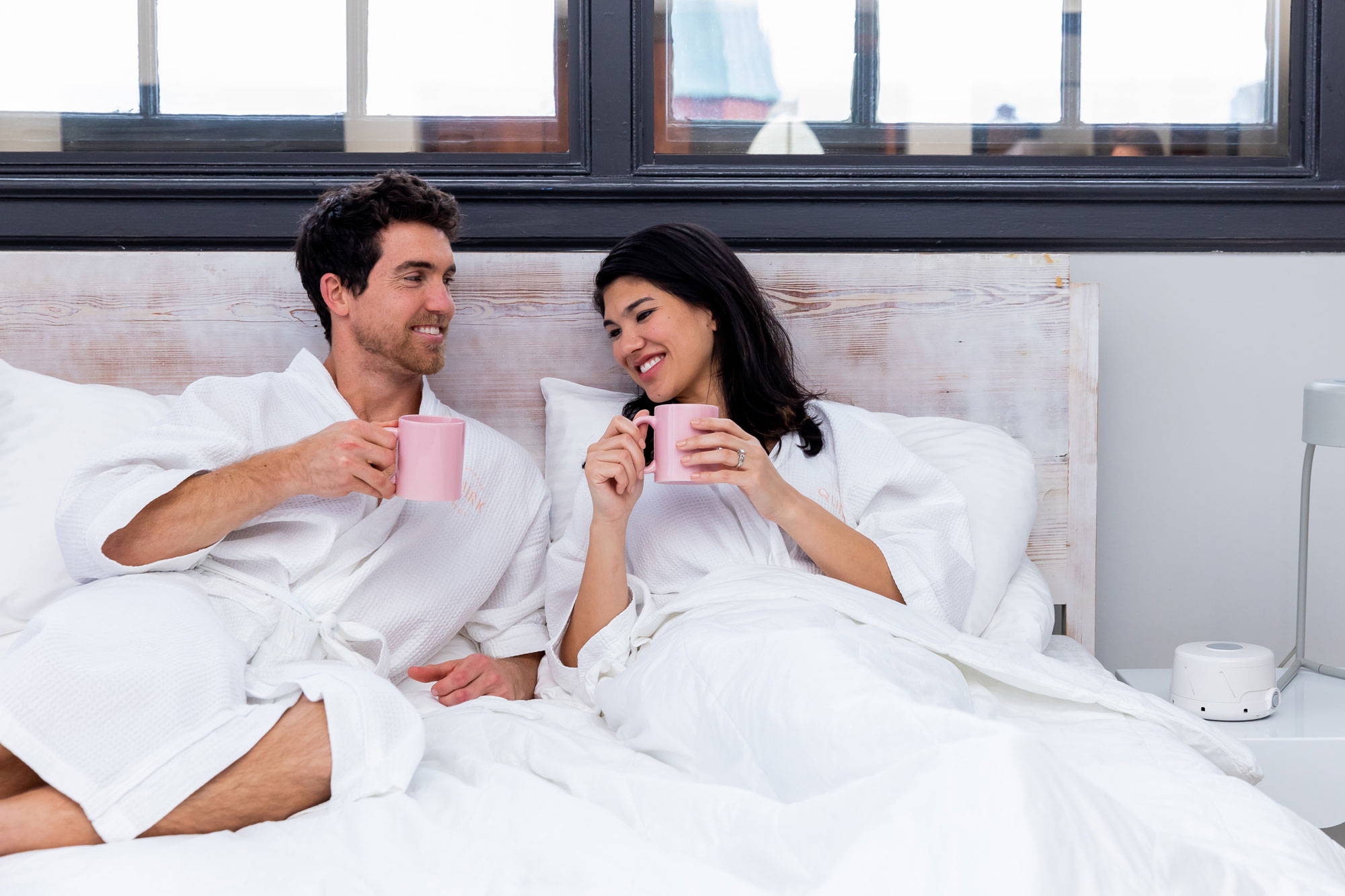 Man and woman in robes sharing coffee in bed with a white noise machine on the bedside table in a bright hotel-style room