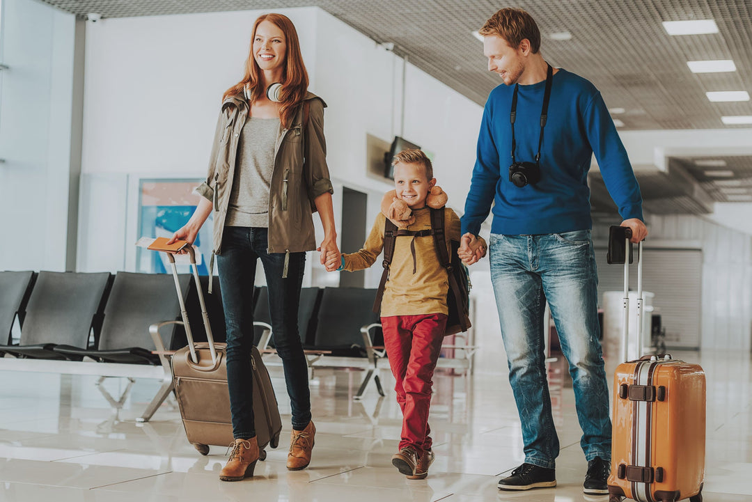 Family walking through an airport terminal holding hands and pulling rolling suitcases; child wears neck pillow