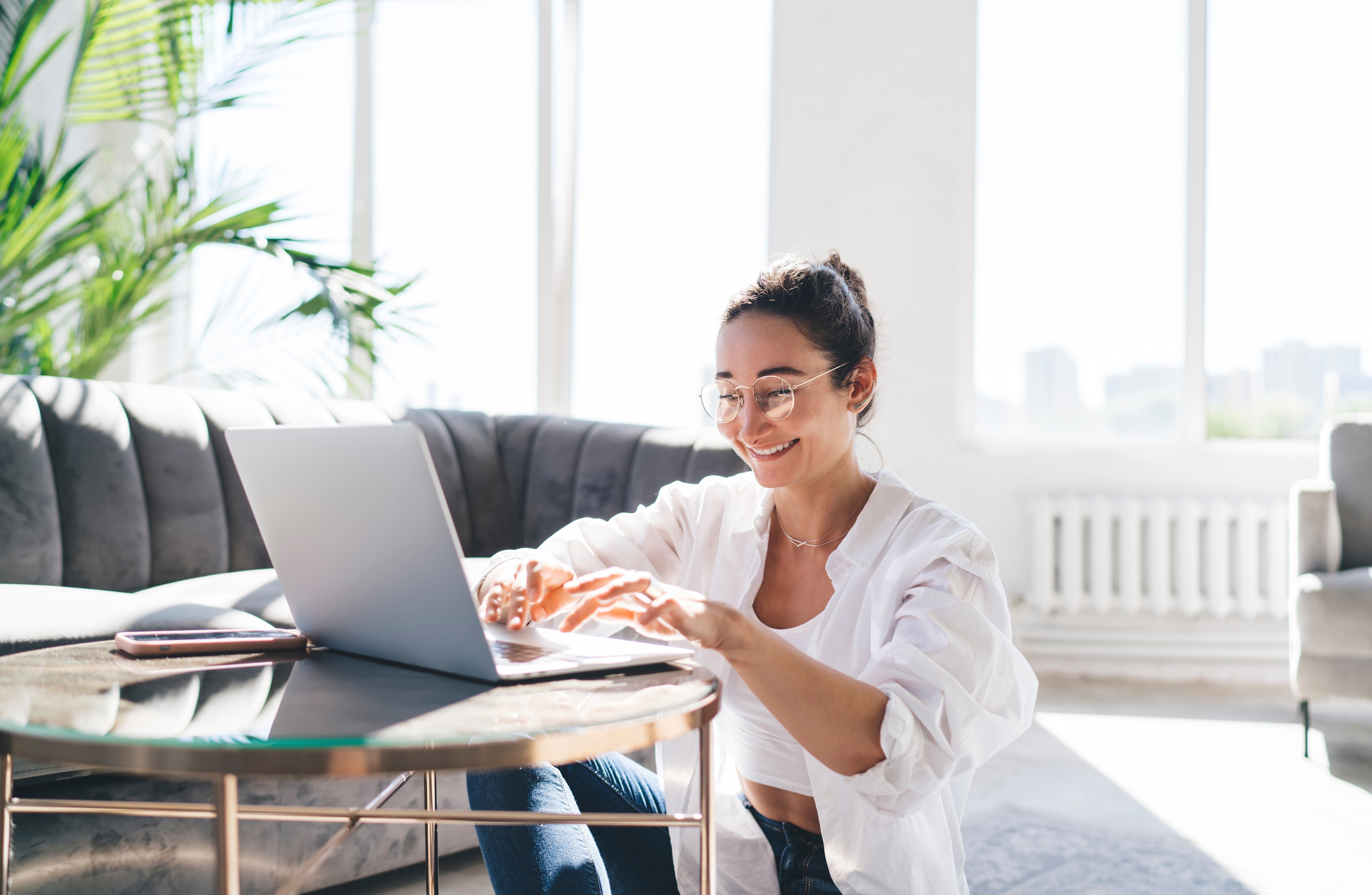 Young woman smiling and typing on a laptop at a sunlit living room table with plants and a city view in the background
