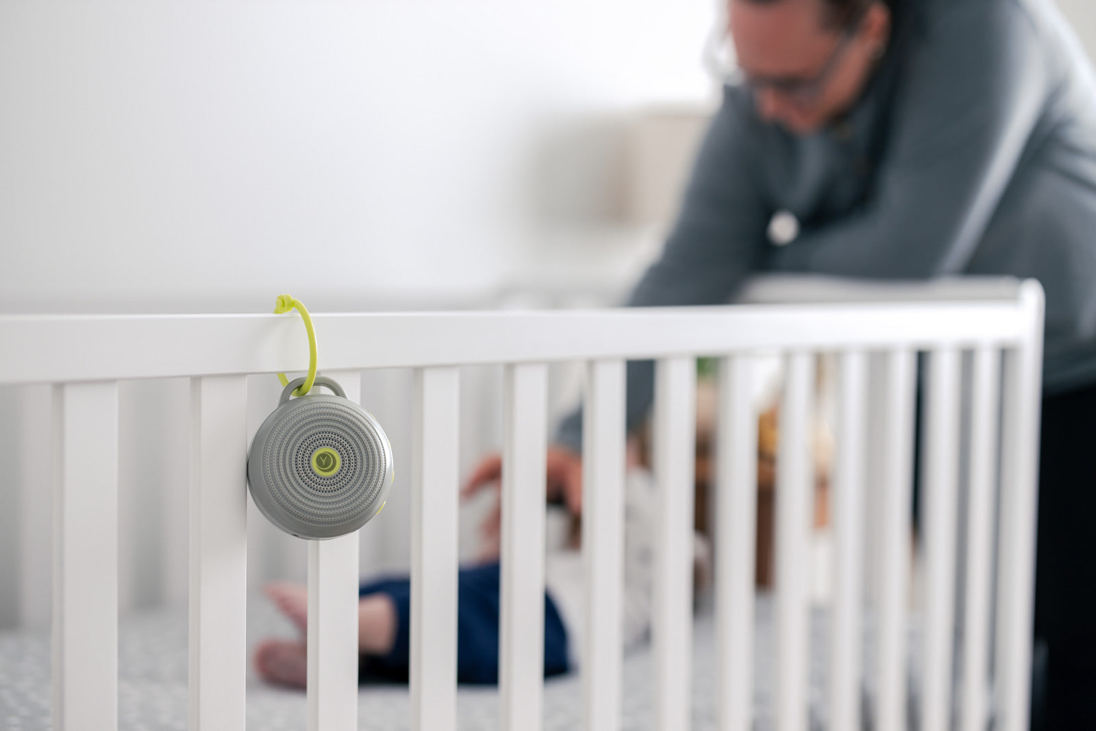 Close-up of compact round white-noise machine hanging on a crib rail with green hook, parent tending baby blurred behind