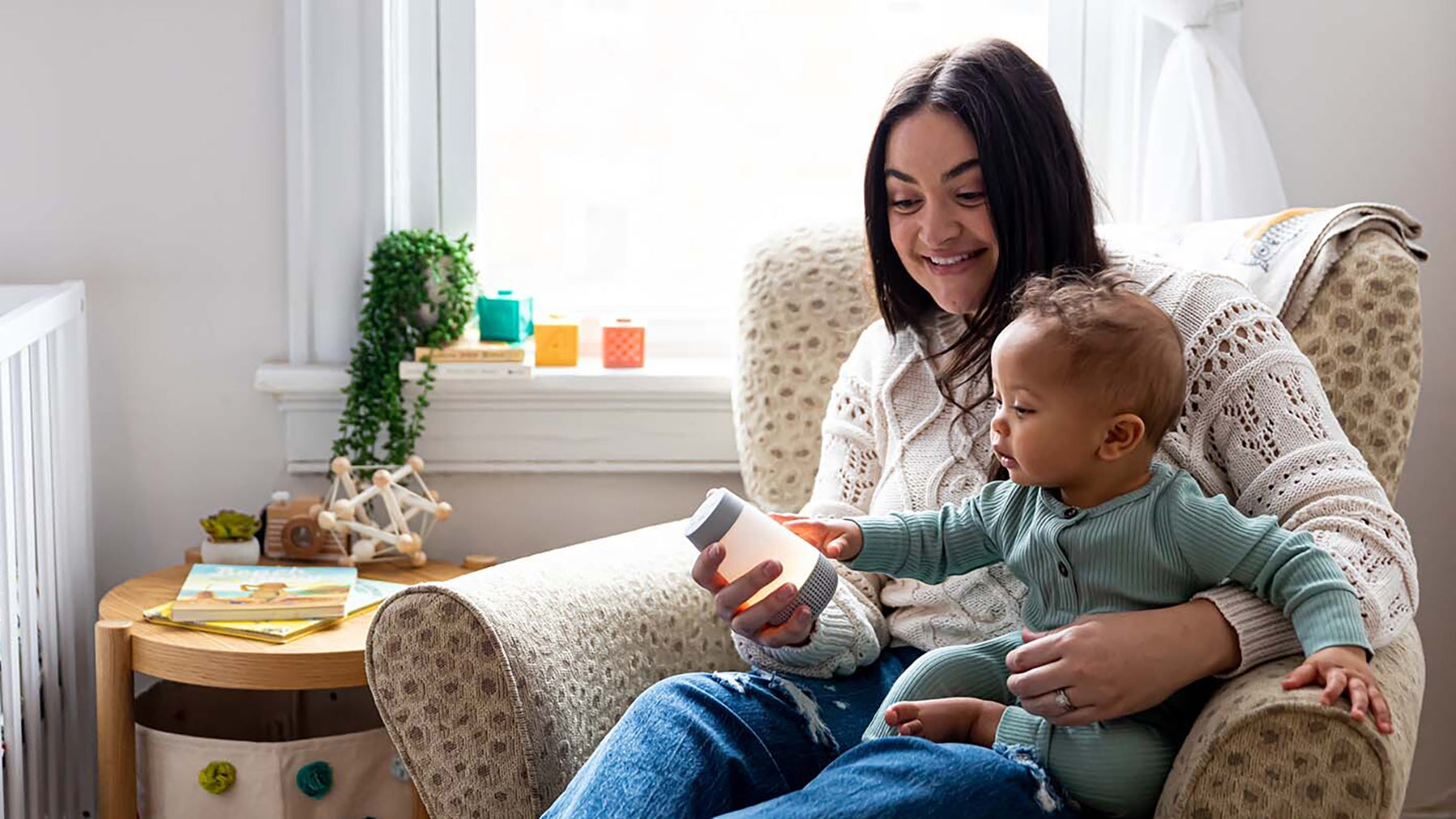 Mother holding a baby who reaches for a glowing white noise machine while seated in a cozy nursery armchair