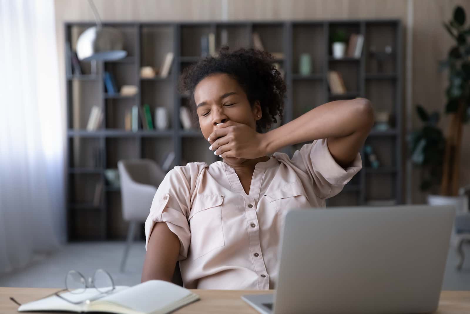 Woman yawning at a desk with laptop and notebook in a home office, looking tired