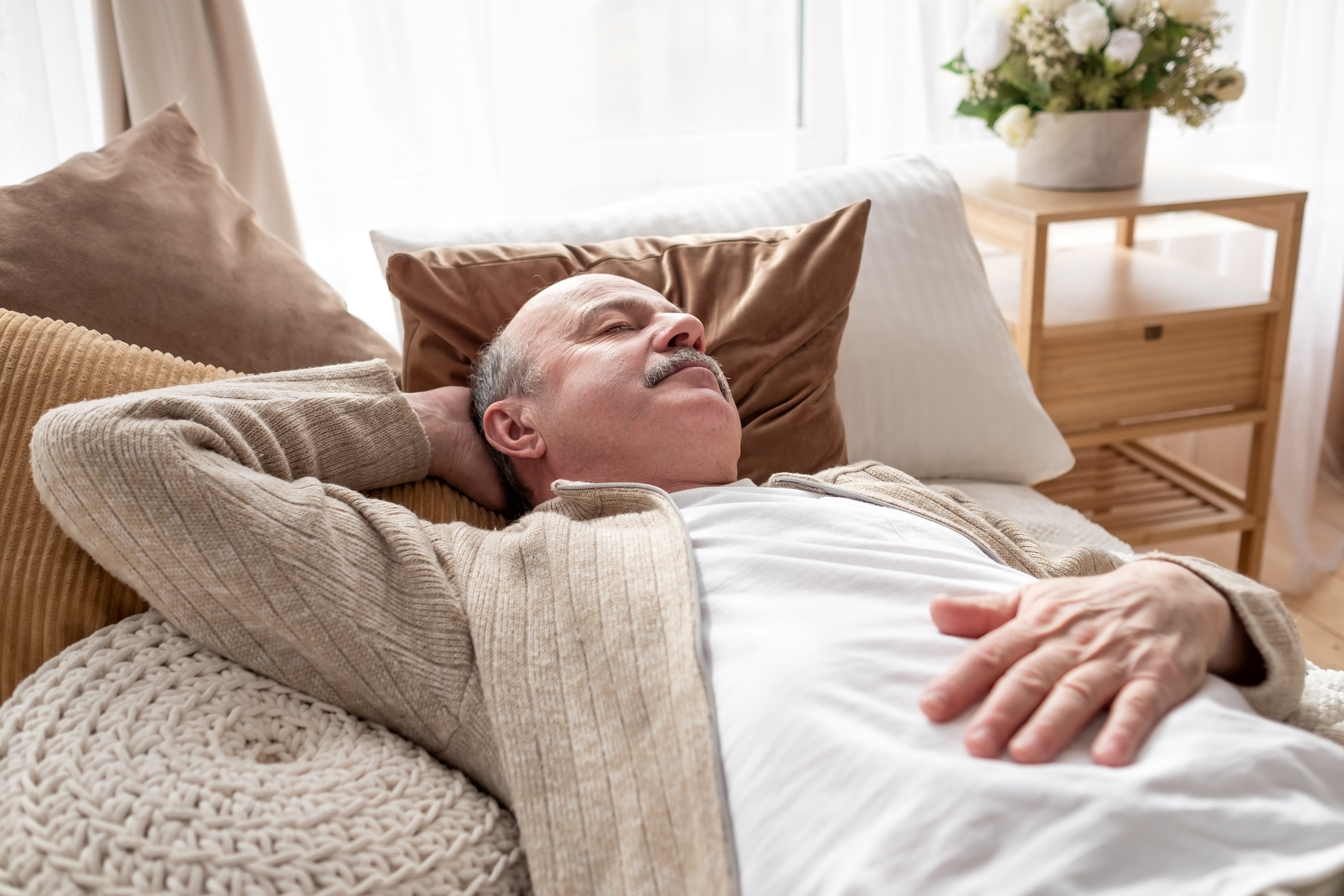Older man resting with eyes closed on a sofa surrounded by pillows in a bright living room