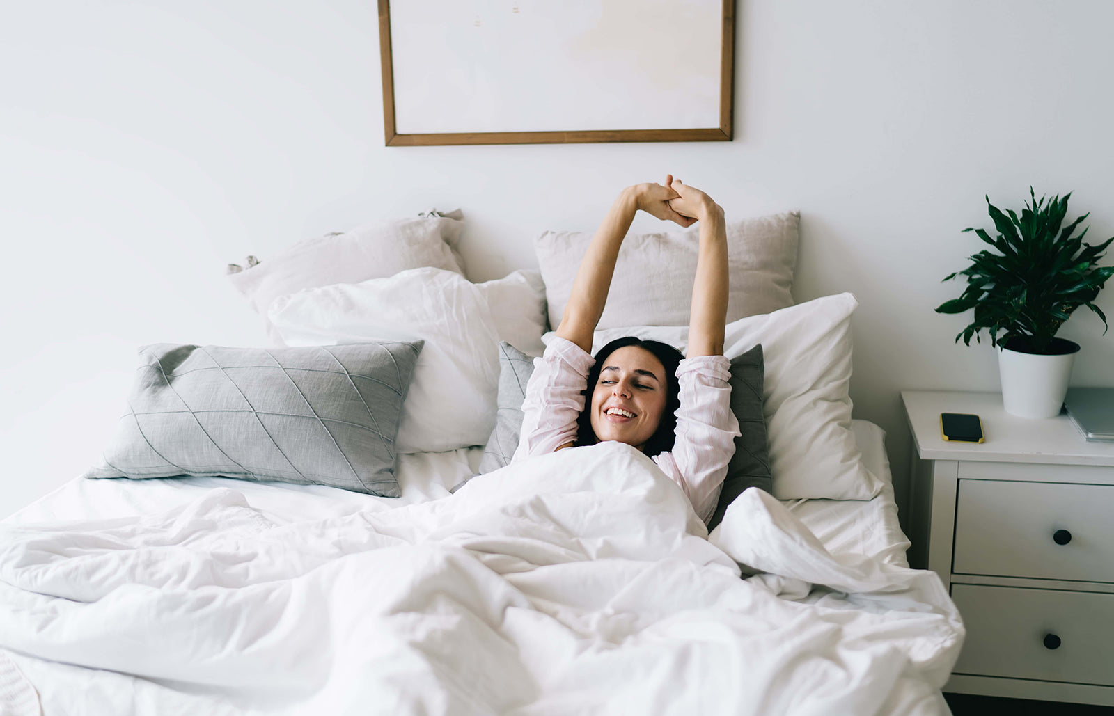 Woman stretching and smiling in bed on white bedding in a bright, minimalist bedroom
