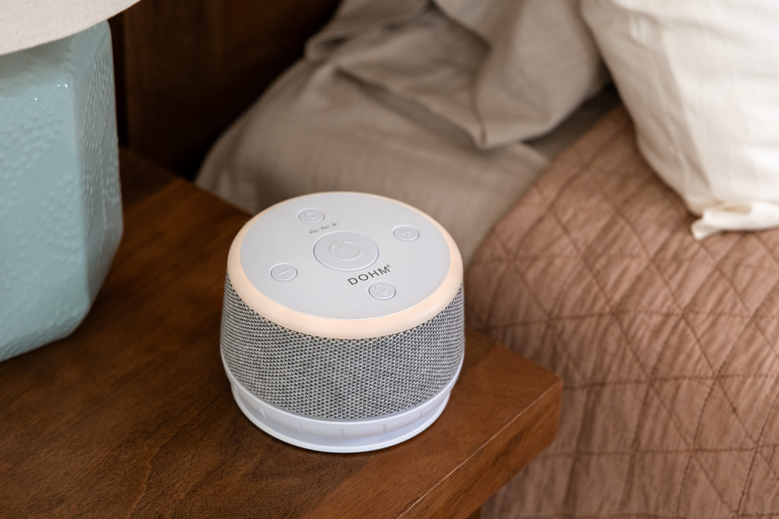 Angled top view of DOHM Nova sound machine showing illuminated ring nightlight and fabric speaker on a wooden bedside table