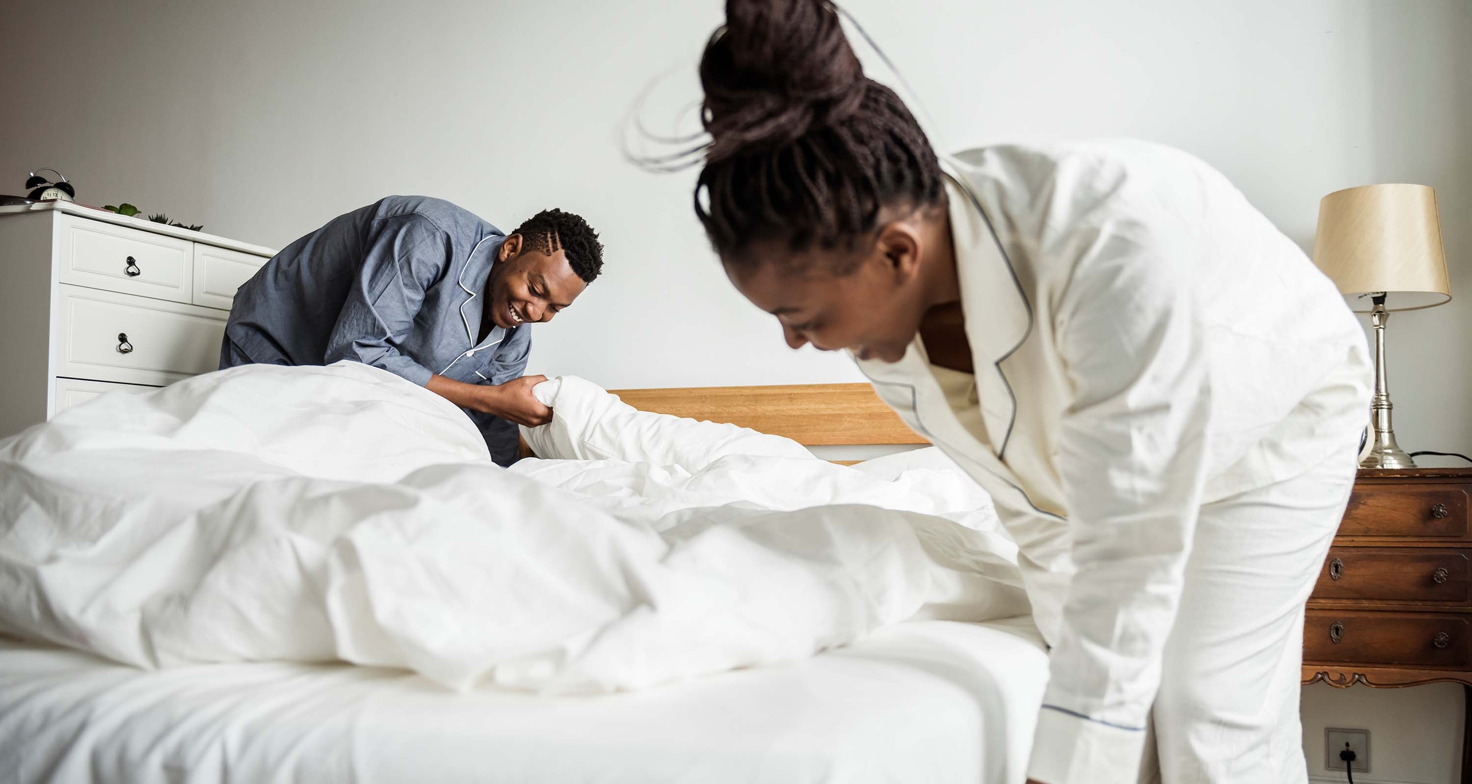 Smiling couple in pajamas making a bed together in a bright bedroom with white bedding and wooden headboard