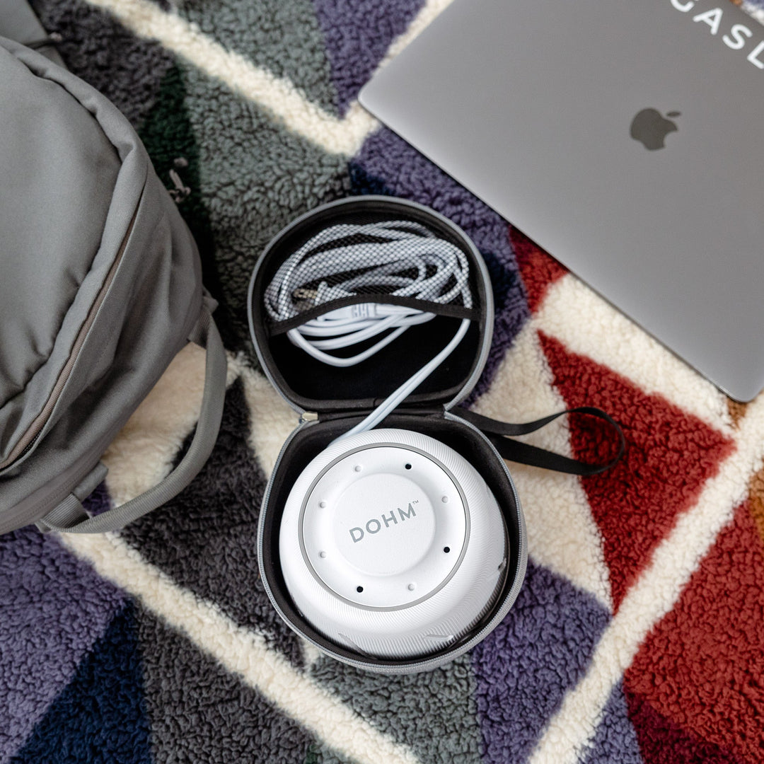 White Dohm sound machine in a travel case beside a laptop and backpack on a patterned rug