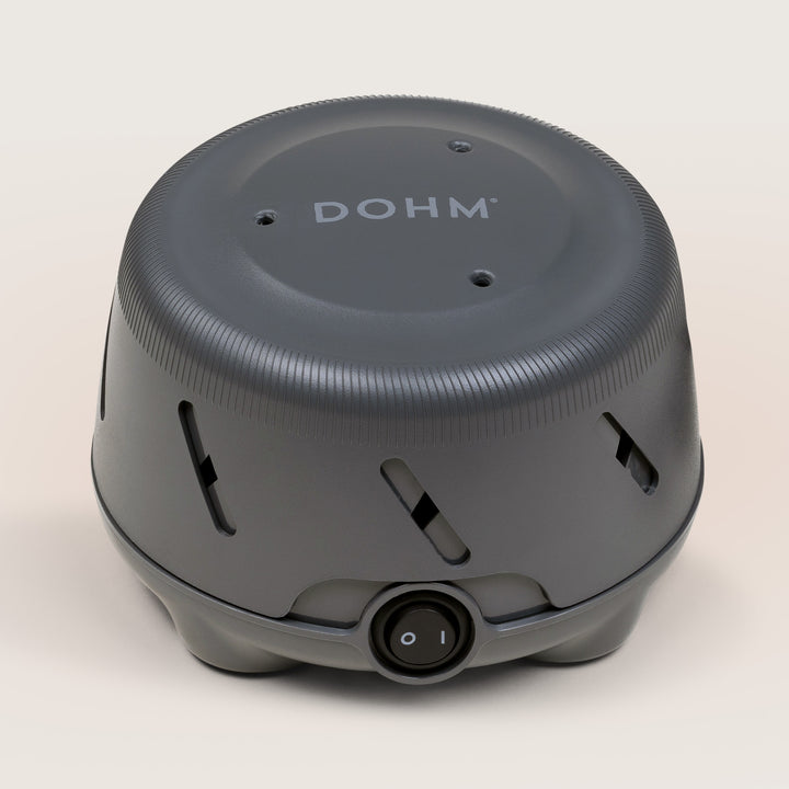 Angled front view of Dohm Uno white noise machine showing power switch, vent slots, and DOHM logo on light linen background